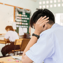 A young boy sits at a school desk with his hand covering his ear