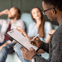 A male therapist wearing glasses takes notes on a clipboard as he counsels a young couple sitting on a couch