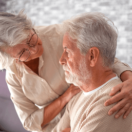 An older woman hugs and comforts a sad looking older man