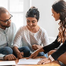 Two young women and one young man wearing business casual clothing are gathered around a table looking at documents together