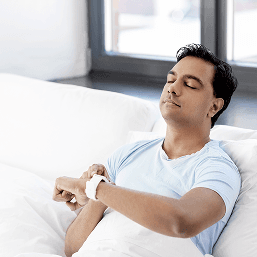A man holds his smartwatch while leaning back on a couch with his eyes closed