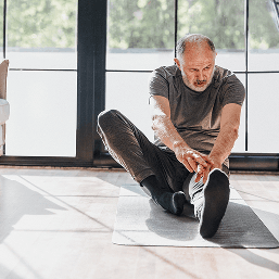 A middle-aged man sits on a yoga mat attempting a yoga pose while looking away